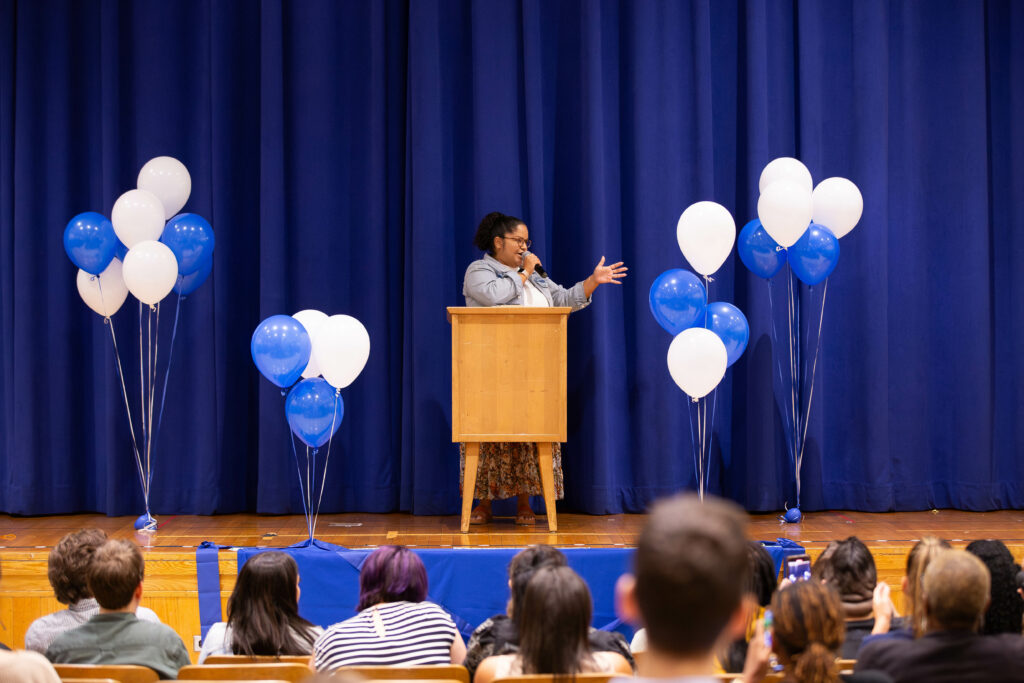 Ms. Sanabria stands behind a wooden lectern on stage in front of a blue curtain with balloons.