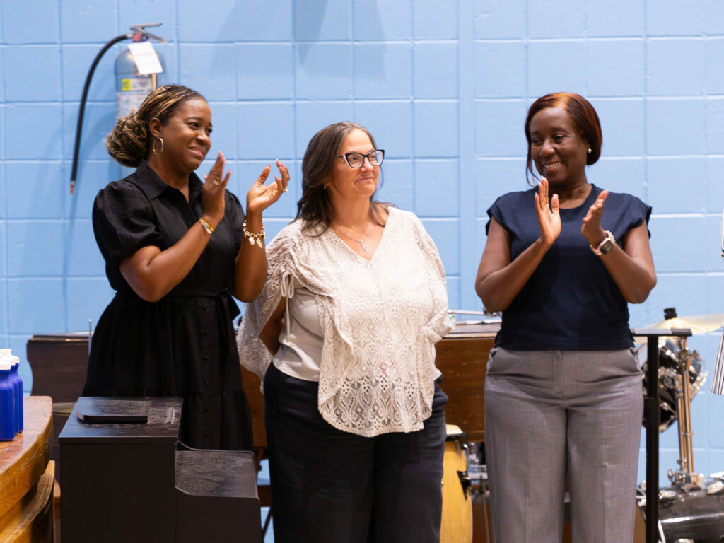 School and district staff clapping in front of a piano with a blue wall in the background.