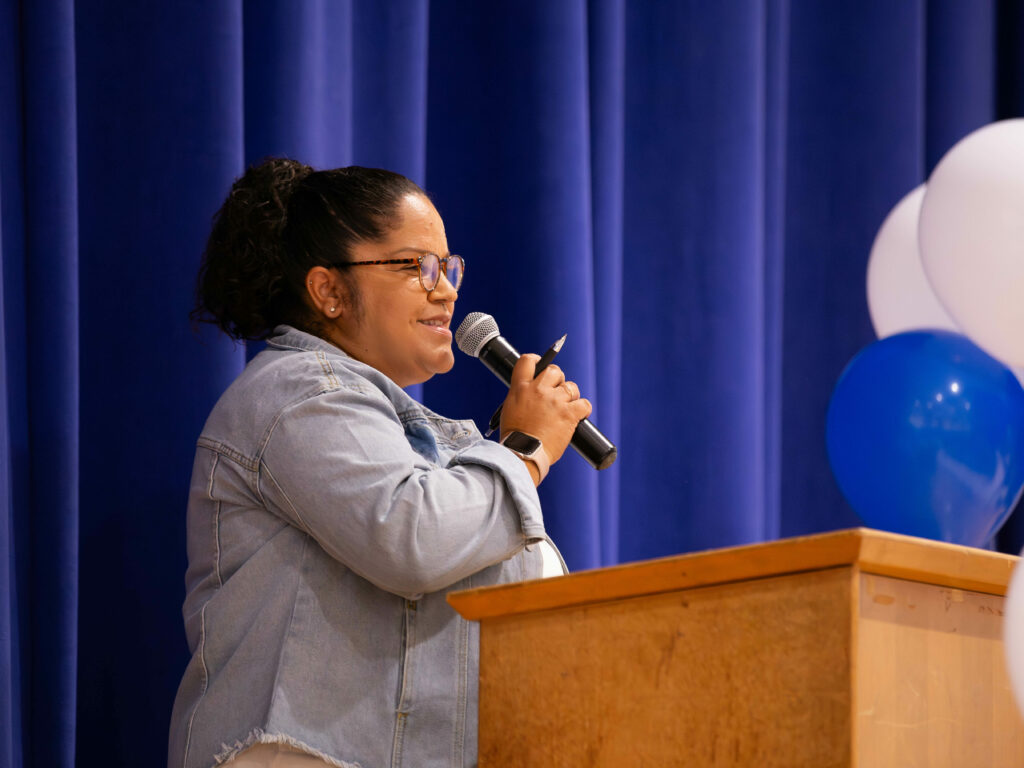 Ms. Sanabria holds a mic and stands behind a lecturn delivering a speech at our IB ceremony