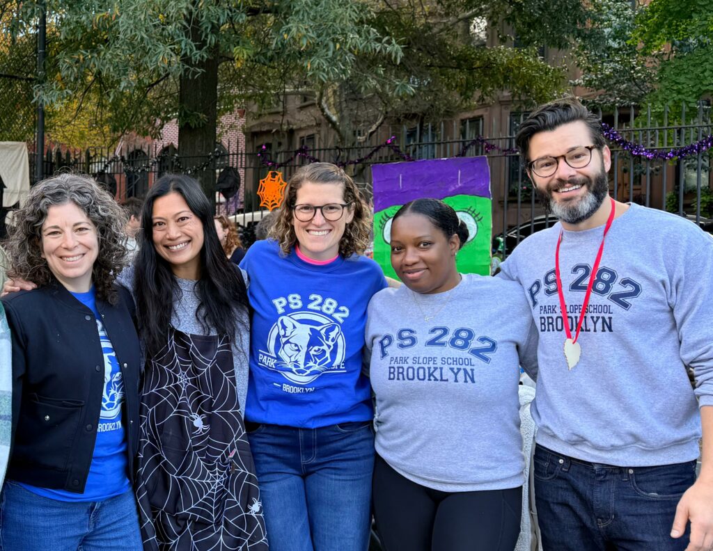Group of parent volunteers wearing PS 282 swag smile outside on the turf