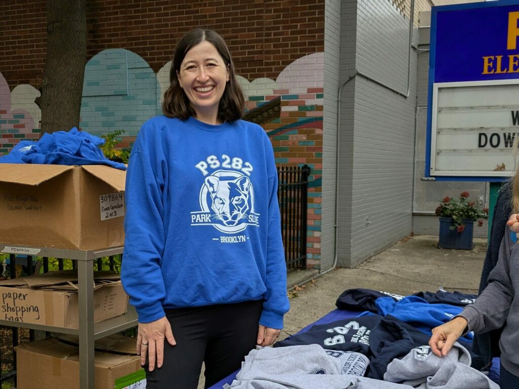 Brigit stands behind a merch table wearing PS 282 blue sweatshirt merch