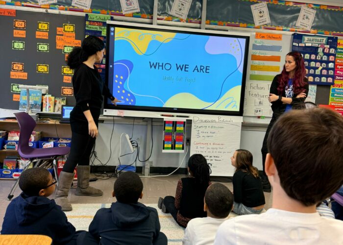 Students sit facing a smartboard with two teachers standing on either side. The slide reads, "Who We Are."