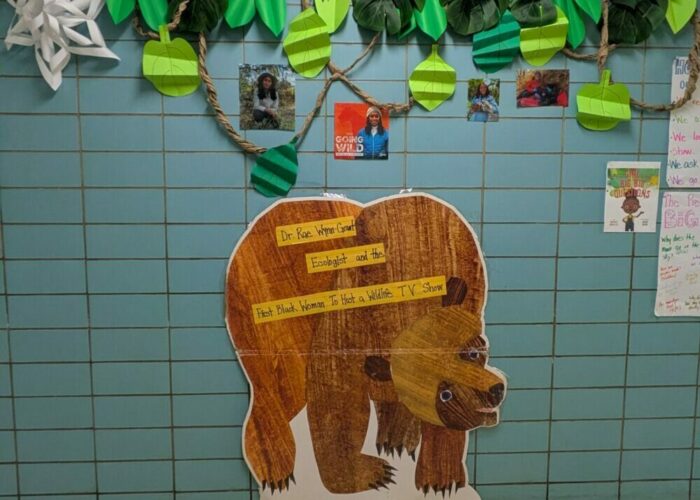 Hallway display with a large paper bear, green paper leaves hanging above, and small photos and student work on a tiled wall.