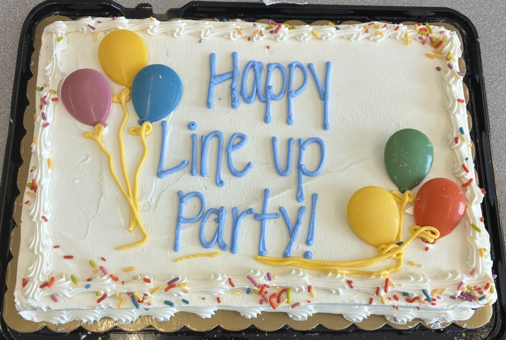 A sheet cake with white frosting, colorful sprinkles, balloon decorations, and blue icing text that reads ‘Happy Line Up Party!’