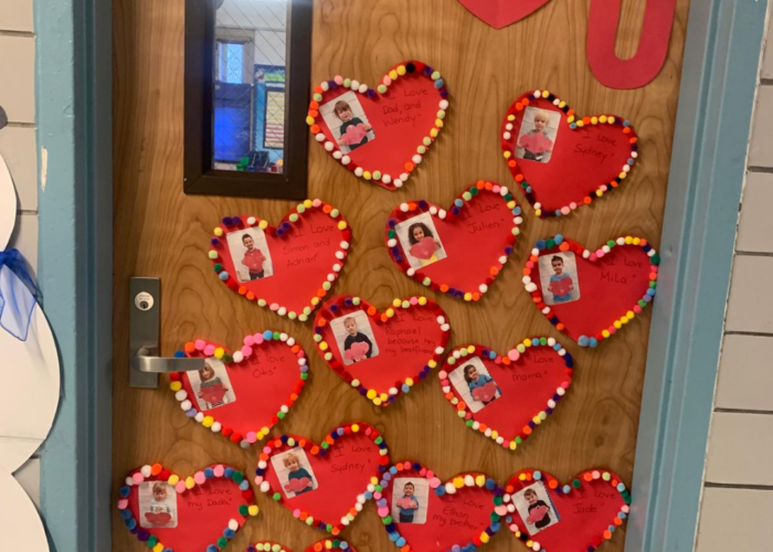 A classroom door decorated with red paper hearts bordered in colorful pom‑poms, each displaying a small photo and writing, with large cutout letters spelling ‘I ❤️ U’ above.
