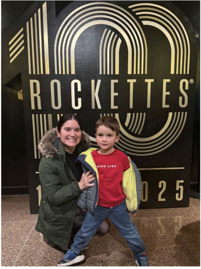 Mom crouches down next to son in front of a black and gold anniversary sign with the words "100 Rockettes" in the background.