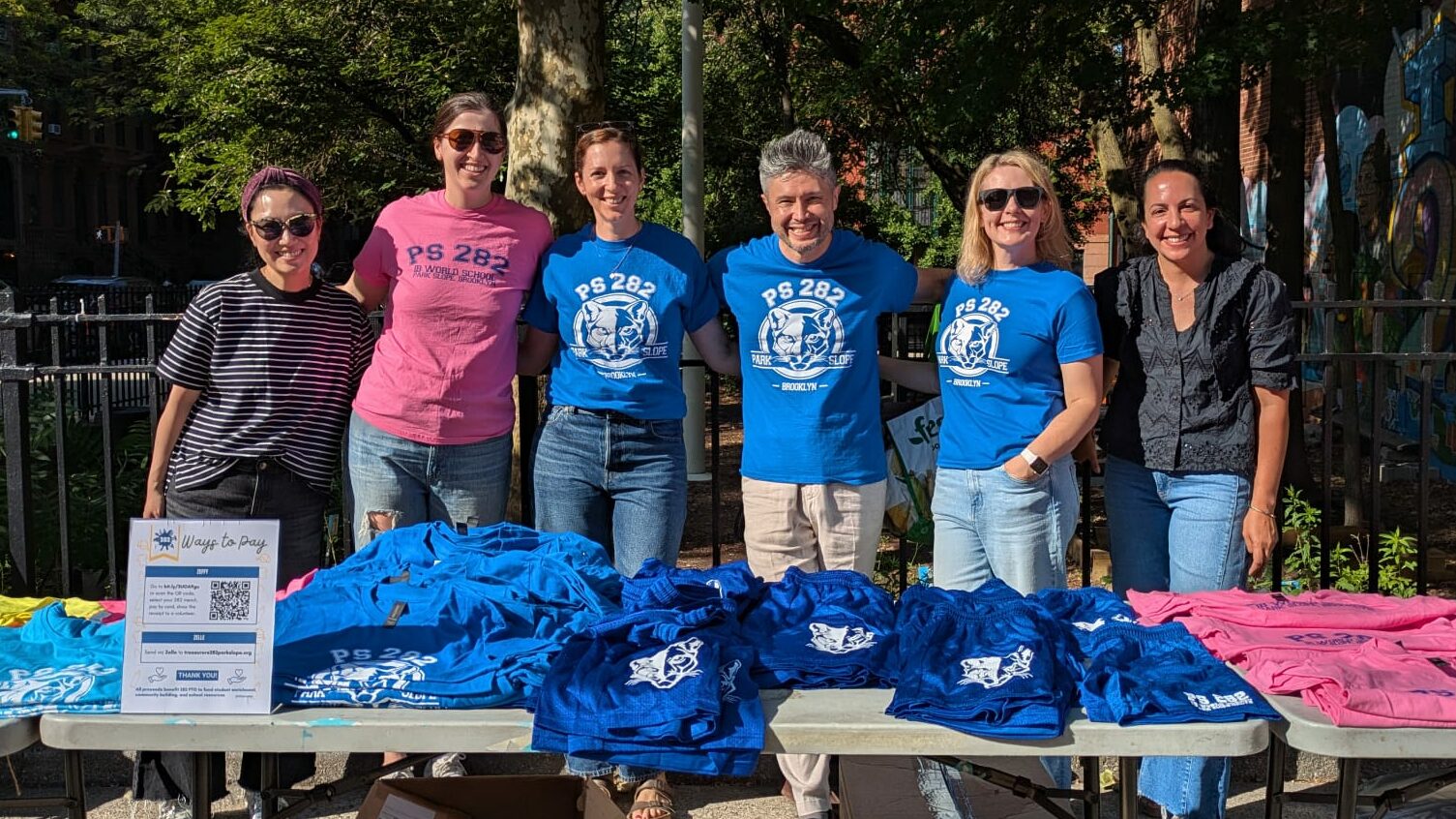 Group of parent volunteers wearing tshirts that read PS 282, standing together behind a table of PS 282 merch with the school garden and fence in the background.